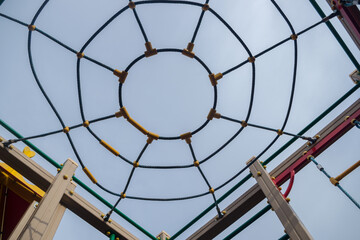 A spider-like installation of a rope dome crowning the top of a structure supported by wooden posts with metal cross bars for climbing children on the playground. View from below against the blue sky.