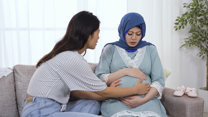 portrait asian female is touching her painful muslim friend’s abdomen gently and trying to help relief her anxiety during contraction.