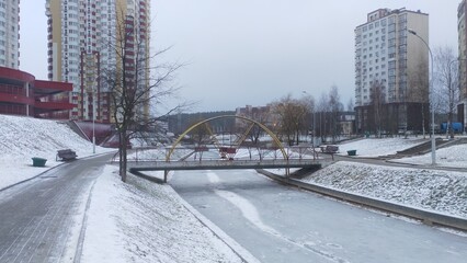 Ice has formed on the canal with water in the city square. The banks where trees, benches and light poles stand are covered with snow. A concrete bridge with a metal fence has been built 