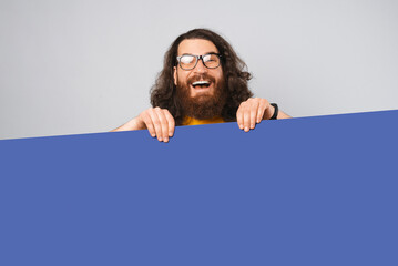 Ecstatic bearded man is hiding behind copy space blue poster in a studio.