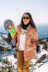 Russia. Sheregesh. Girl snowboarder in black sun goggles in winter in sunny weather outdoors among the rock and snow. The girl is standing with a snowboard in her hands, smiling and posing. Sport