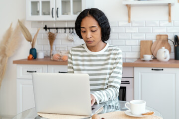 African-american female using laptop while sitting in kitchen. Confident and focused on remote work