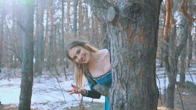 Fantasy happy medieval woman playing hide and seek, peeking out from behind tree, smile on face, beckoning hand gesture follow me. Girl princess in winter forest, bright sun. Blue vintage velvet dress