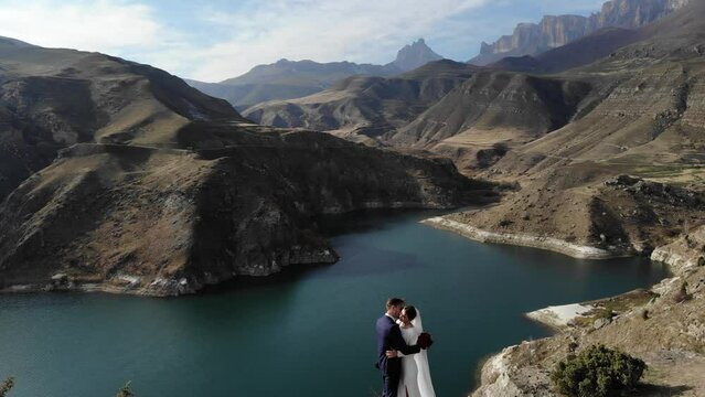 Aerial view from a couple in love standing on the edge of a cliff near the water of a mountain lake. A woman in an amazing white dress and a man in a suit. People newlyweds on a wedding walk