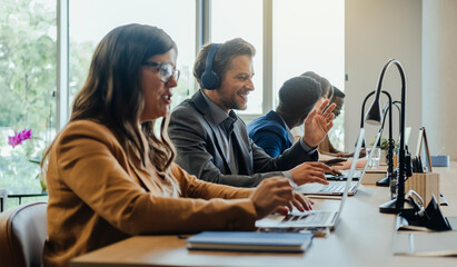 Happy Business People Sitting in a Row and Working on Computers in Open Plan Office. 
Five colleagues typing something on a laptop computers and talking on a video call with a clients.