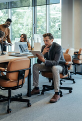 Serious Business Man Using Digital Tablet at Work. 
Businessman sitting in open plan office and reading business report on a tablet while his multi-ethnic team of colleagues talking in the background.