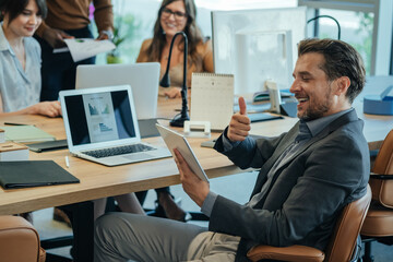 Happy Businessman Showing Thumbs Up to Digital Tablet at Online Meeting with Client.

Smiling business man talking on a video call on his tablet while sitting in open plan office desk with colleagues.