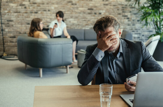 Disappointed Business Man Using Mobile Phone at Work. 

Overworked displeased businessman holding hand over his face after receiving bad news on his laptop computer or phone in open plan office.