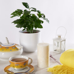 Still life with a standing candle, a cup of tea and a teapot. White wooden table and light background.