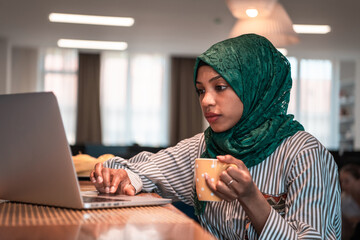 African muslim business woman wearing a green hijab drinking tea while working on laptop computer in relaxation area at modern open plan startup office.