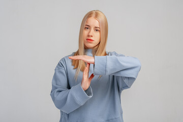 Confused blonde woman makes pause or break time gesture, timeout sign, asks to wait, upset about deadline, stands against grey background. Body language concept. Time limit