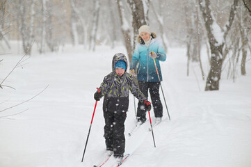 A boy with a woman learns to ski while walking in a snowfall. Outdoor activities for children in winter. Physical education lessons. - 488979973