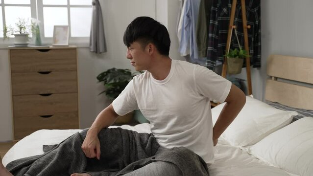 side view of a stressed asian man suffering lower back pain is pounding massaging himself to ease discomfort after rising in the morning on bed at home