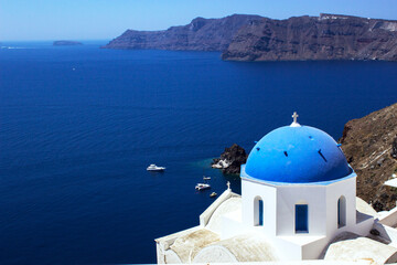 Fototapeta na wymiar Church of Santorini with its traditional blue dome with the sea and cliffs in the background and boats anchored in a small harbor, travel concept.