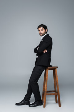 full length view of elegant man in black suit posing with crossed arms near high stool on grey