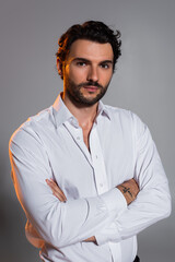 Obraz premium bearded man in white shirt standing with crossed arms and looking at camera on grey
