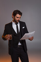 man in black elegant suit reading newspaper while holding glass of whiskey on grey background