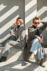 Two stylish beautiful young women in fashionable clothes with sunglasses are sitting outside near a concrete wall at sunlights. Women's retro urban style and fashion