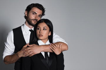 elegant man hugging brunette asian woman and looking at camera on grey background