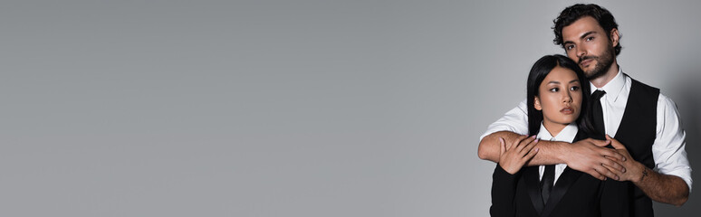 elegant man looking at camera and hugging young asian woman on grey background, banner