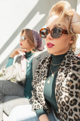 Obraz premium Fashion blonde woman with sunglasses in stylish leopard coat and sweater sit on the street with girlfriend. Urban female style autumn clothes