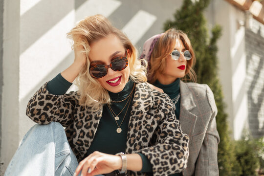 Image of two young happy women friends with fashion sunglasses in trendy vintage urban clothes sits over gray wall with sunlights. Looking at camera