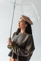 positive asian woman in leather pullover and beret looking away under transparent umbrella isolated...