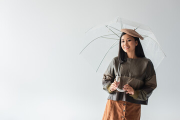 smiling asian woman in leather pullover and beret posing under transparent umbrella isolated on grey