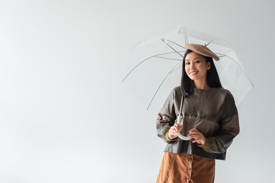 smiling asian woman in leather pullover and beret posing under transparent umbrella isolated on grey
