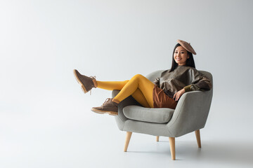 full length view of happy asian woman in leather pullover and yellow tights sitting in armchair on...