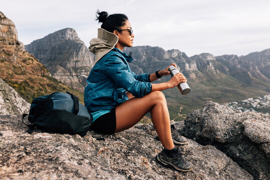 Side view of woman hiker in sports clothes enjoying the view. Female in sunglasses holding a thermos sitting on a rock and relaxing during a hike. - Powered by Adobe
