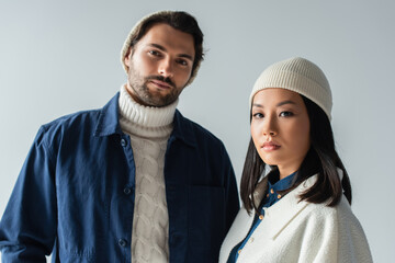 multiethnic couple in stylish clothes and beanies looking at camera isolated on grey