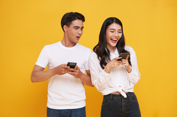 Shocked asian man spying on her smiling girlfriend while both using mobile phones isolated over yellow background. chatting or social media concept.