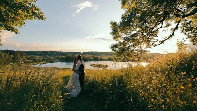 Side view of attractive bride and groom standing, hugging on field in setting sun rays. Camera approaching to wedding couple while they kissing with beautiful lake on background. Concept of wedding