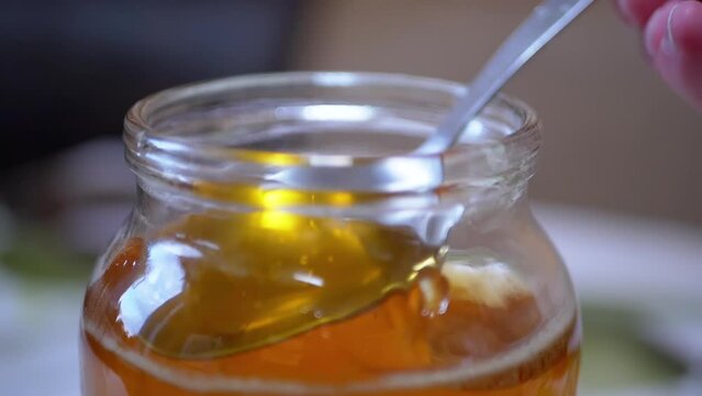A Female Hand Picks Up with a Spoon Transparent, Liquid Honey from a Glass Jar. Yellow thick flow bee honey pouring, falling smoothly stream in tank. Healing, organic beekeeping product. Zoom.
