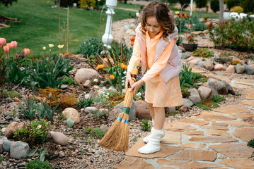 
A little girl with the help of a broom sweeps the floor in the dacha in the garden. Landscape....
