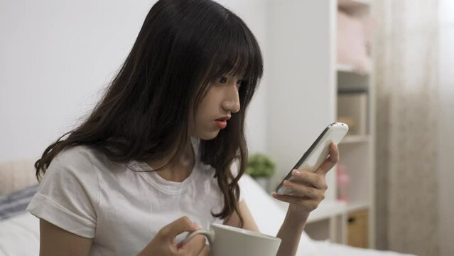 closeup shot of a shocked asian girl carrying tea cup is scrolling down on her smartphone screen to check a surprising online news in the bedroom at home.