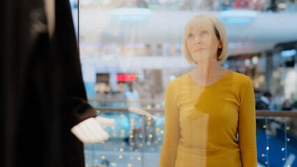 Mature middle aged caucasian woman looking sadly at showcase window clothing store in shopping mall upset frustrated sad because higher prices costly purchase luxury too expensive not affordable