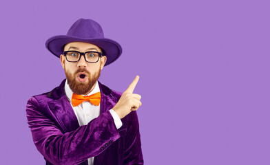 Man with surprised expression on his face points his index finger at copy space on purple background. Funny man in purple velvet jacket and hat shows place for your text or promotional product.