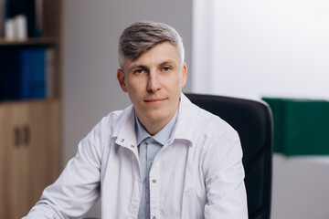 Portrait of male doctor physician in medical uniform. Family therapist sitting at workplace desk, looking at camera.