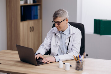 Male doctor wearing glasses and uniform working typing on computer. Family therapist consulting patients online through digital health platform.