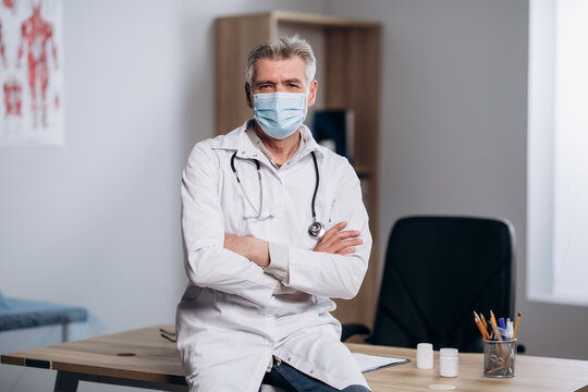 Portrait of male doctor in medical mask posing for camera with arms crossed. Family therapist sitting at workplace.