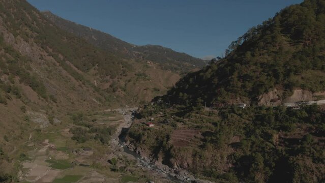remote farm town in mountainous tropical region Kabayan Benguet Philippines aerial fast approaching wide angle over river on edge of valley blue sky sunny day