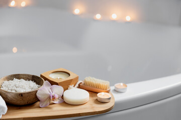 Fototapeta na wymiar Preparation for hotel spa treatment, home bath procedure. White washbasin in bathroom, accessories on tray. Burning candles, soap, foot brush, towel, glass bottle with sea salt, orchid flower