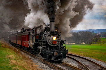 Obraz na płótnie Canvas An Antique Steam Passenger Train Traveling Thru Farmlands Puffing Lots of Smoke on a Cloudy Winter Day