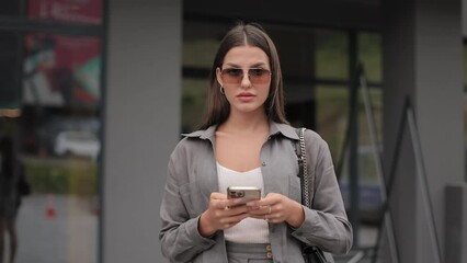 Adorable young woman looking at camera with confidence in eyes while standing on street and holding mobile in hands. Charming lady wearing trendy sunglasses and stylish clothes.