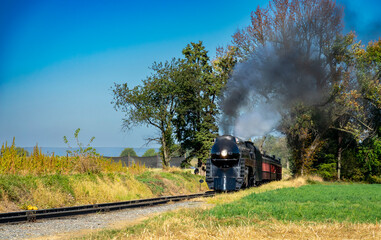 Obraz premium An Antique Restored Steam Engine Approaching Head-on, On Some Old Rail Road Tracks Blowing Smoke and Steam Traveling thru Farmlands.