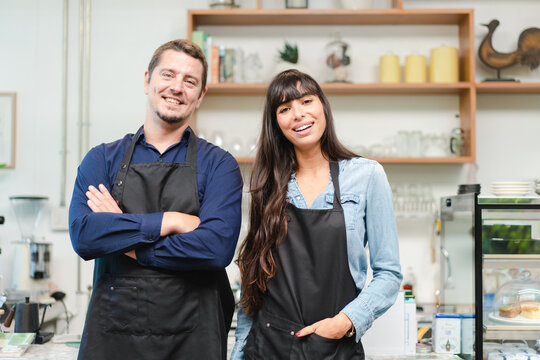 Portrait of couple barista owners in apron smiling and standing on front counter in coffee shop