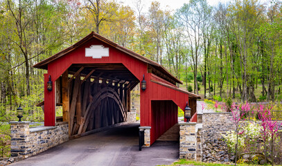 Fototapeta na wymiar View of a Restored Burr Truss Covered Bridge on a Country Road With Stone Approach Walls on a Mostly Sunny Day