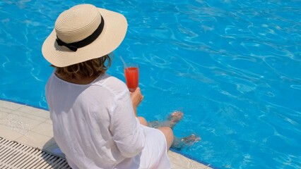 woman in a straw hat relaxes near the swimming pool, drinks a pink cocktail and dangles her legs in the water. Summer mood concept